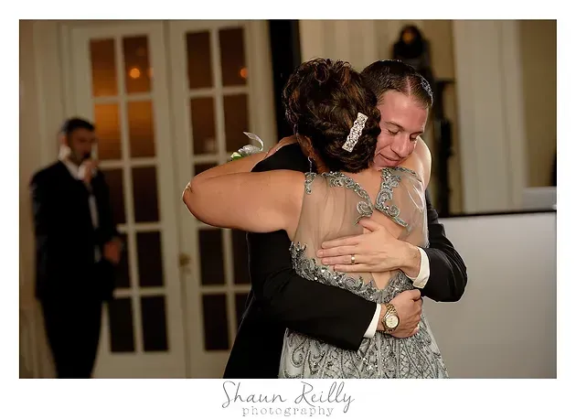 Two people hugging at a formal indoor event, smiling amid soft warm lighting.