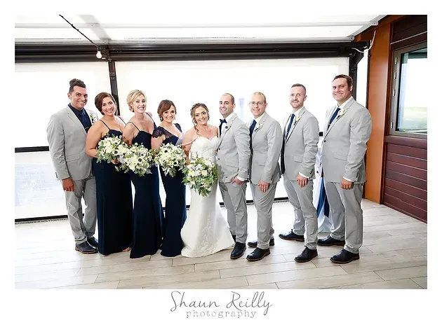 Wedding party posing outdoors in formal attire, with bridesmaids holding bouquets and groomsmen in gray suits