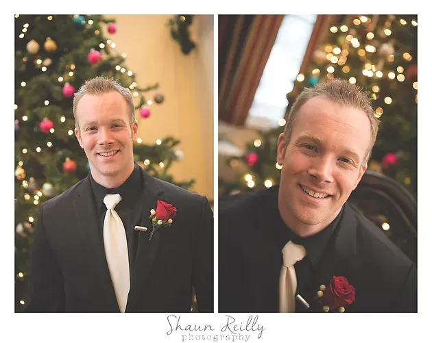 Man in black suit with white tie and red boutonniere, posing in front of a lit Christmas tree.