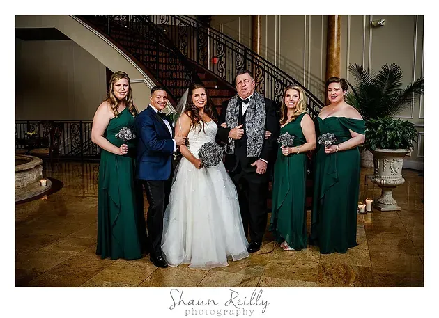 Wedding party posing indoors, bride in white gown with attendants in green dresses and one man in blue suit.