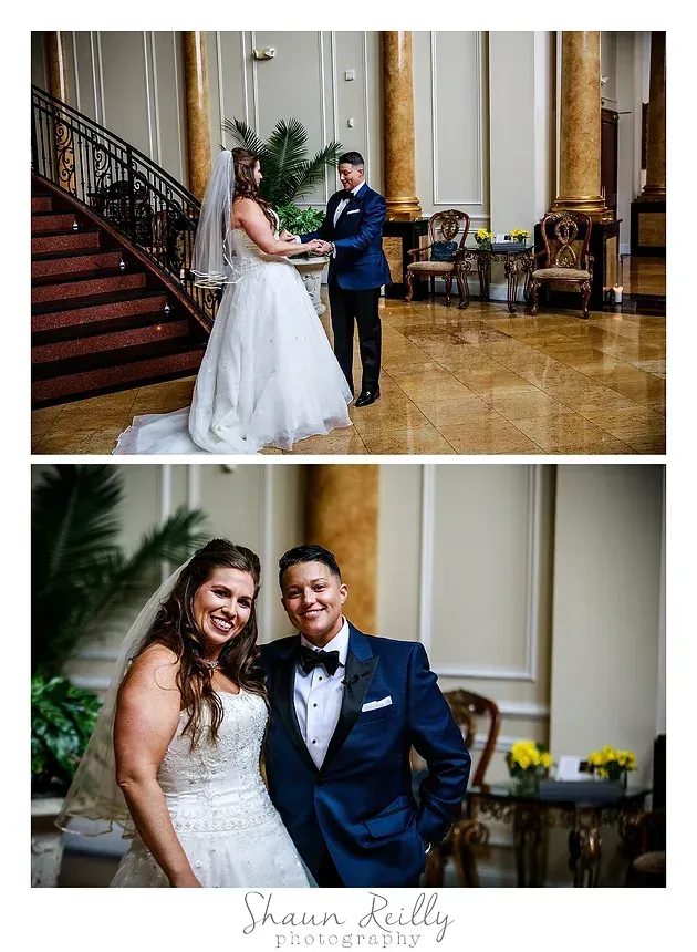 Bride and groom pose in a grand ballroom beside a staircase, with ornate columns and polished marble floors.