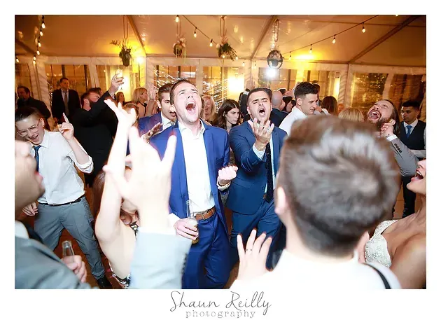 Guests dancing at a lively indoor reception under string lights, with a cheering couple centered.