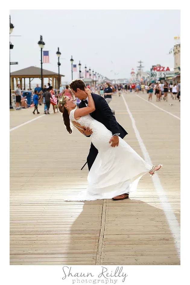 Wedding couple kissing and dipping on a boardwalk by the beach, with spectators and colorful lamp posts nearby