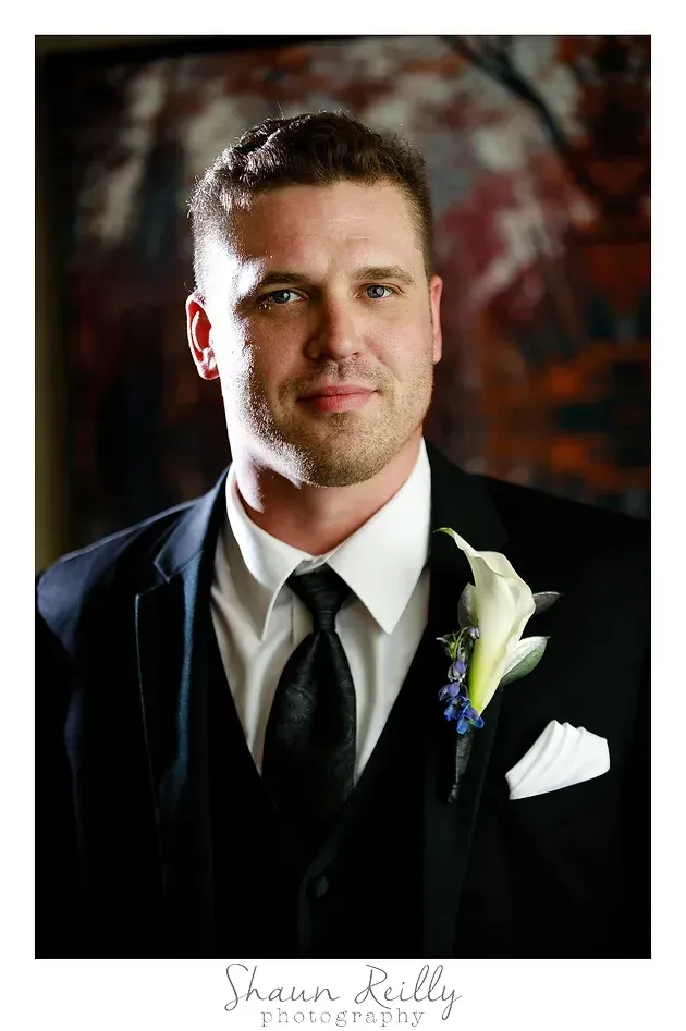 Man in black suit and tie with white boutonniere, posing in a formal portrait against a dark background
