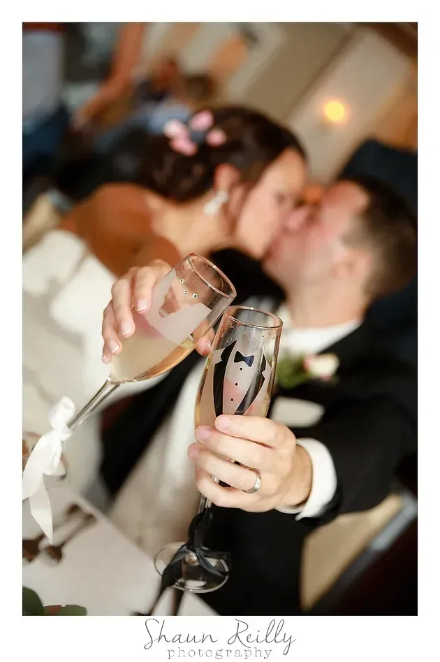 Clinking champagne flutes at a wedding, with a blurred couple kissing in the background
