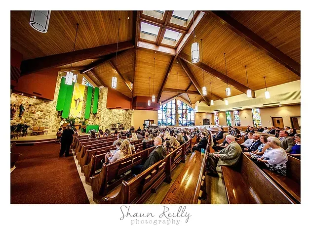 Church congregation seated in wooden pews under a high timber ceiling, facing the altar and stained glass.