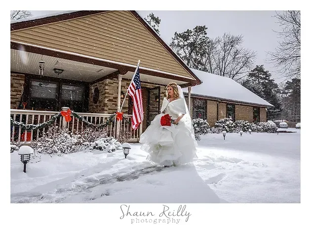 Bride in white gown holding red flowers beside a snow-covered house with an American flag