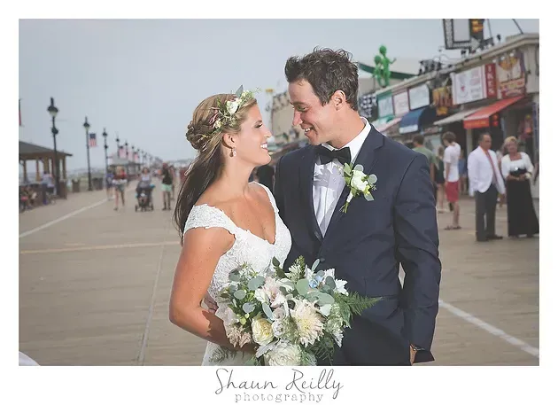 Newlyweds smiling on a seaside boardwalk, bride holding bouquet and groom in navy suit