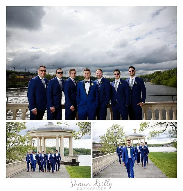 Five men in blue suits posing outdoors by a waterfront and gazebo under cloudy skies