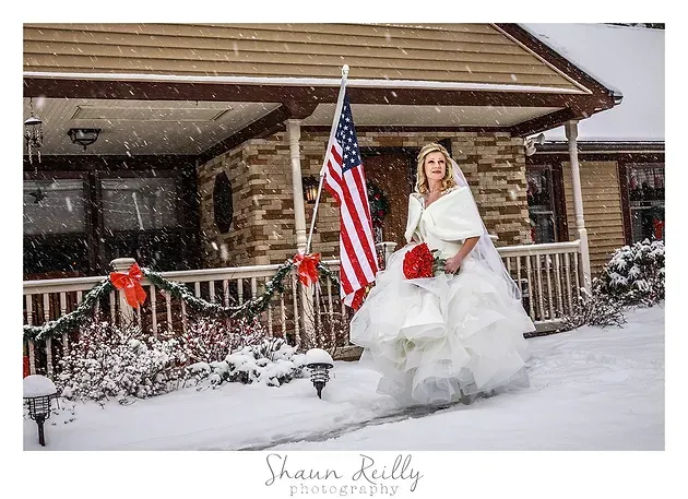 Bride in a white gown holding a bouquet beside an American flag in front of a snowy house