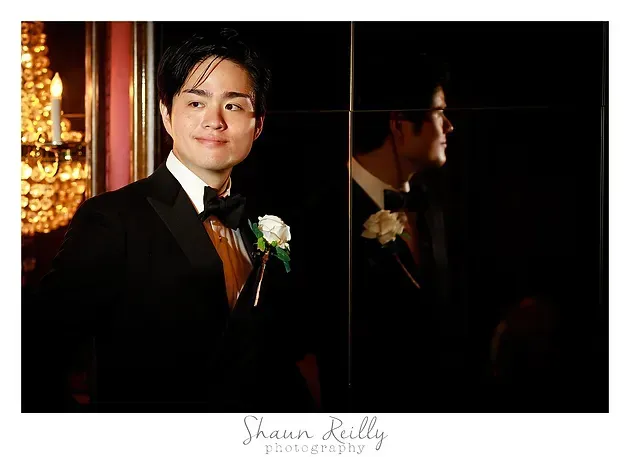 Man in a black suit with a white boutonniere stands beside a mirror in a dimly lit room