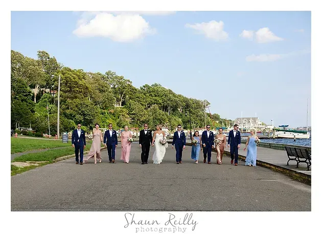 Group in formal attire walking along a waterfront path beside trees, boats, and a blue sky