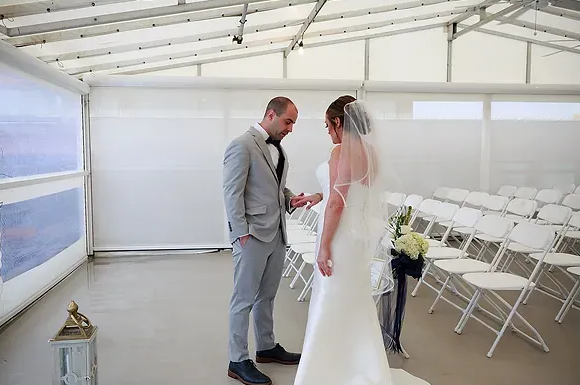 Bride and groom standing together in a white wedding tent, facing each other near rows of chairs.