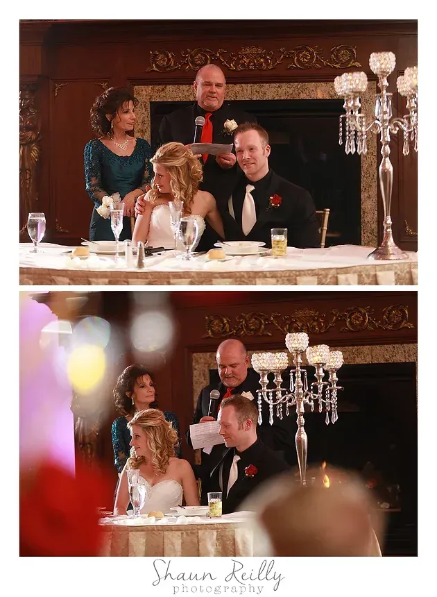 Wedding couple seated at a reception table, with guests standing behind them and crystal decor nearby.