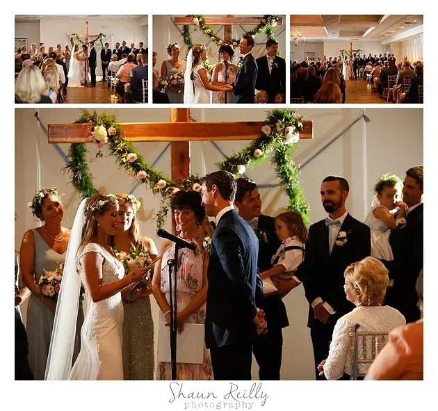 Wedding ceremony under a flower-draped cross, with the couple exchanging vows before seated guests.