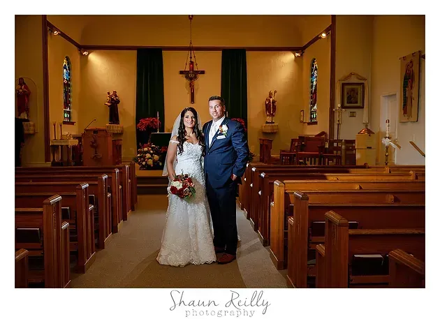 Bride and groom standing in a church aisle with wooden pews and a crucifix behind them