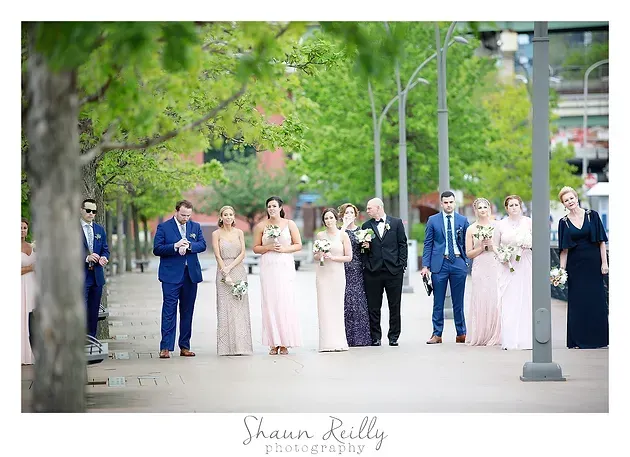 Wedding party lined up outdoors on a tree-lined street, wearing pastel formal attire.