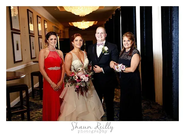Four people posing indoors at a wedding, including the bride in a white gown holding a bouquet.