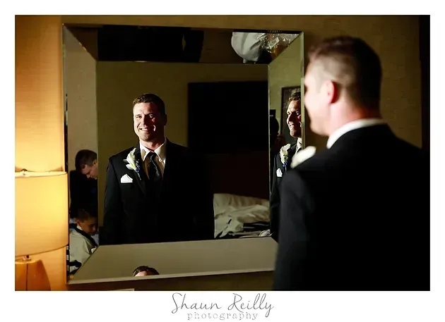 Man in a tuxedo smiling into a mirror, seen beside his reflection in a dimly lit room