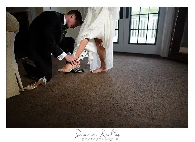 Man kneeling to fasten white dress shoes on a woman in a white dress indoors by a window