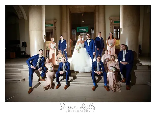 Wedding party posing in front of columns, with bride in a white gown centered among guests in blue and pink attire
