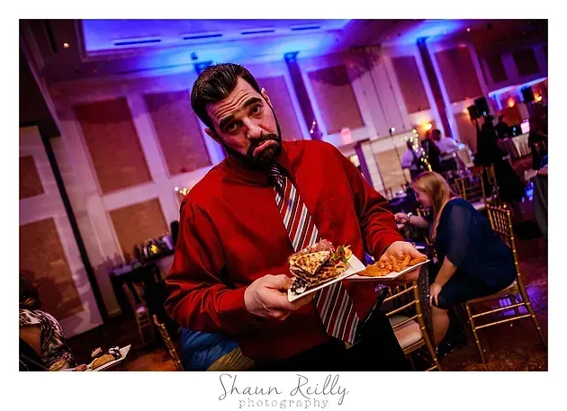 Man in a red shirt holding a plate of food at a banquet hall, smiling at the camera.