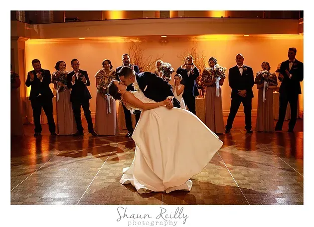 Bride in white gown dancing with partner in a warmly lit ballroom, guests watching in the background