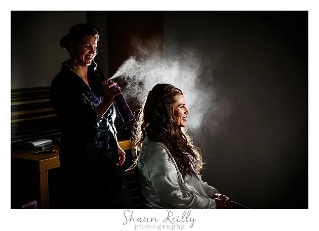 Woman styling another woman’s hair in a dark studio, with spray and soft light.