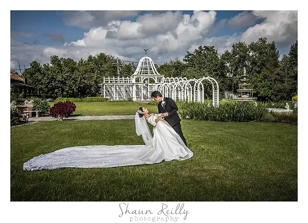 Bride and groom posing in a garden before a white gazebo, with the bride's long veil trailing on the grass