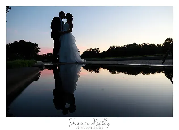 Silhouetted couple embracing at sunset beside a reflective pool, with a wedding dress visible