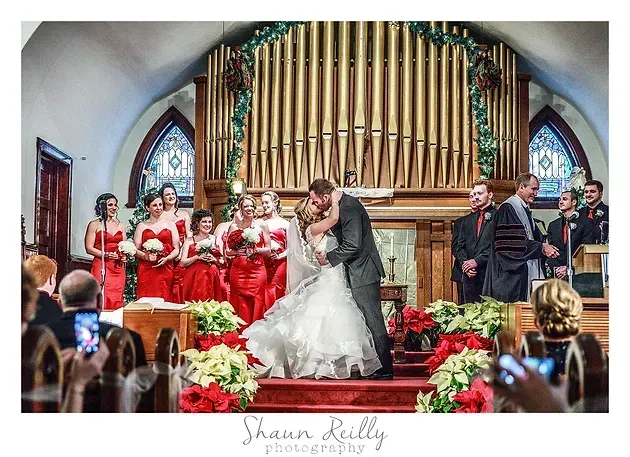 Wedding couple kissing at altar with bridesmaids in red dresses and groomsmen in suits in church