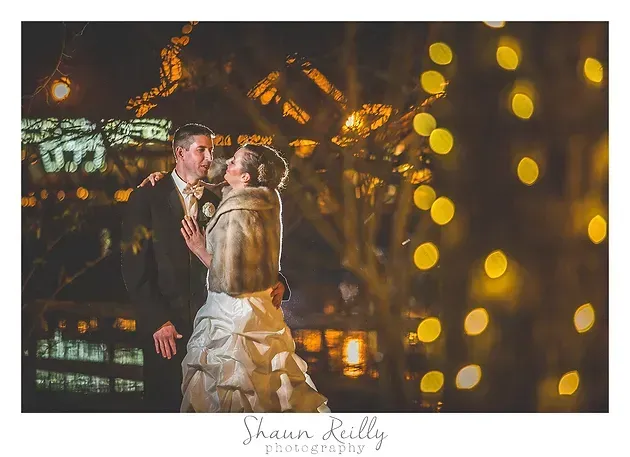 Bride and groom kiss at night with warm city lights and golden bokeh in the background