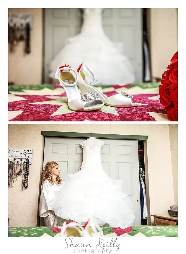 White bridal shoes on a red floral bedspread beside a white wedding dress being held up indoors