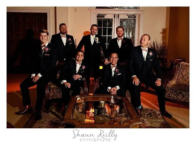 Seven people in suits posing in a formal living room around a glass coffee table.