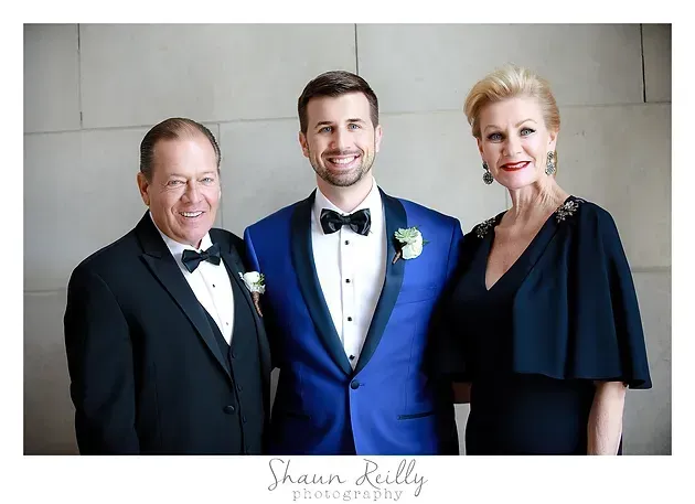 Three people in formal attire posing together indoors, with the center person in a blue tuxedo jacket.