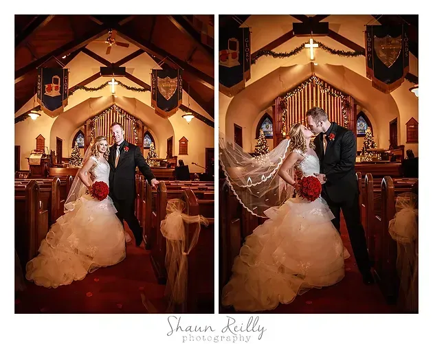 Wedding couple posing in a warmly lit church, bride holding red bouquet and veil flowing.