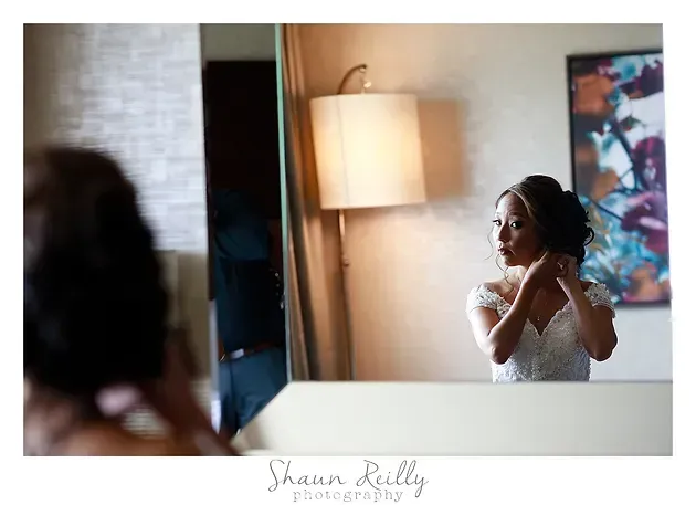 Bride adjusting earrings in a mirror beside a lit floor lamp.