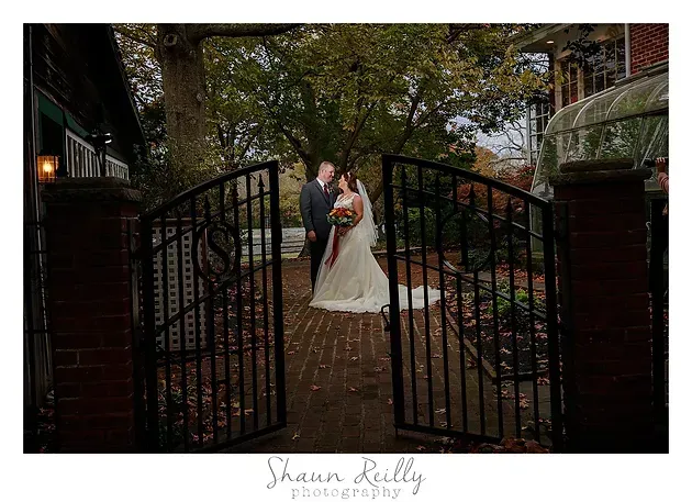 Bride and groom kissing beneath trees by open wrought-iron gates on a garden path