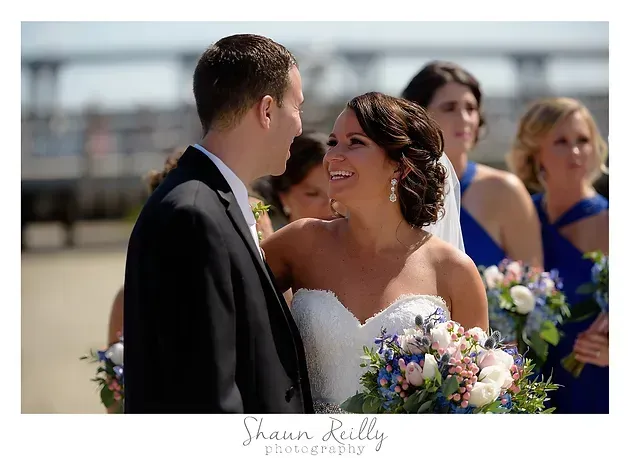 Bride and groom smiling at each other outdoors, with bridesmaids in blue holding bouquets in the background
