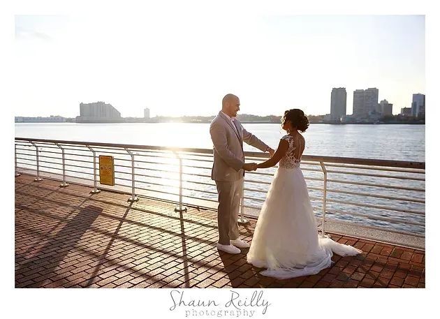 Bride and groom holding hands on a waterfront boardwalk at sunset with city skyline in the background
