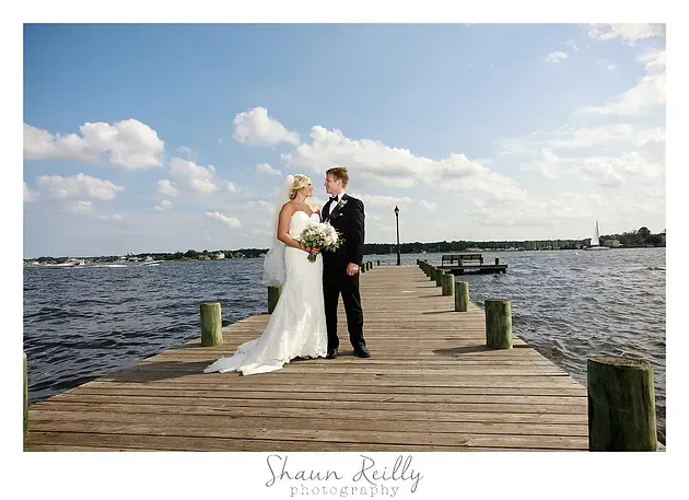 Bride and groom pose on a wooden dock by the water under a partly cloudy sky.