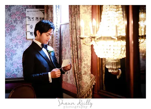 Man in a tuxedo reading a card beside a glowing chandelier in an elegant room