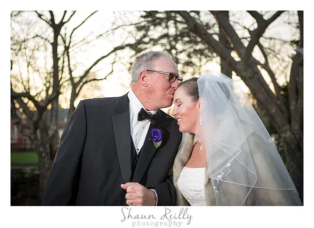 Bride and groom share a kiss outdoors beneath bare trees at sunset.
