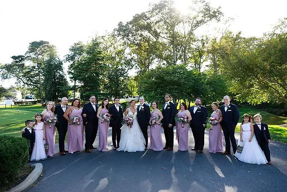 Wedding party posing outdoors on a tree-lined road, with bridesmaids in blush and black and the bride in white.