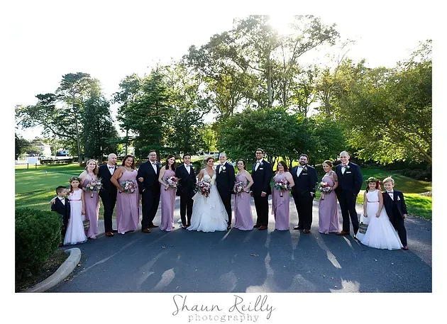 Wedding party posing outdoors on a driveway, with bridesmaids in blush dresses and bouquets, groomsmen in tuxedos.