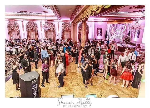 Crowded ballroom dance floor with guests in formal attire under pink and gold lighting
