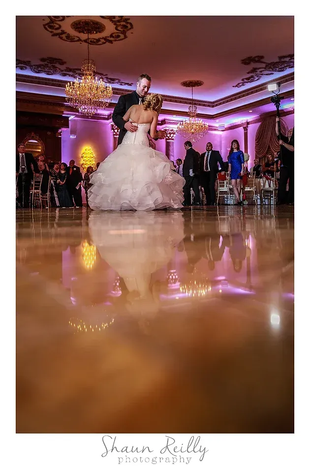 Bride and groom dancing under chandeliers in a warmly lit ballroom, reflected on the polished floor
