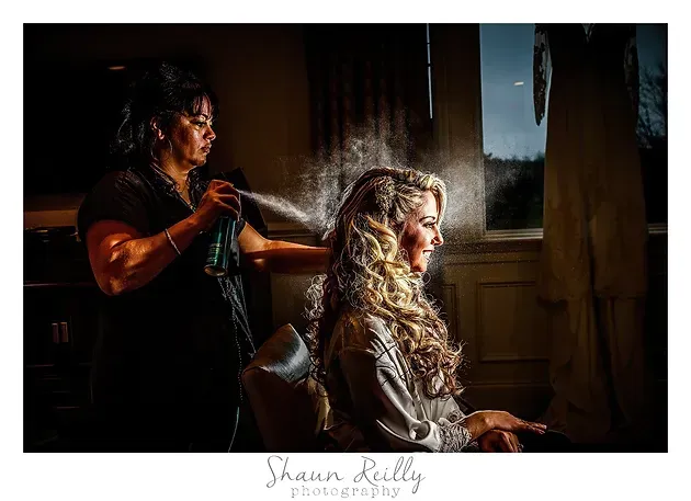 Stylist spraying a woman's blonde hair in a sunlit salon near a window