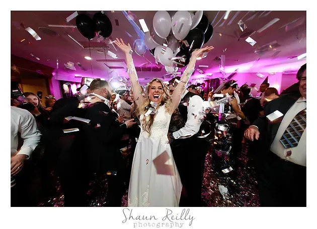 Bride celebrates with guests on a dance floor under pink lights, arms raised amid confetti and balloons.