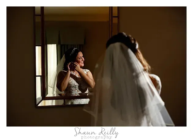 Bride adjusting earrings in mirror while veil drapes in foreground